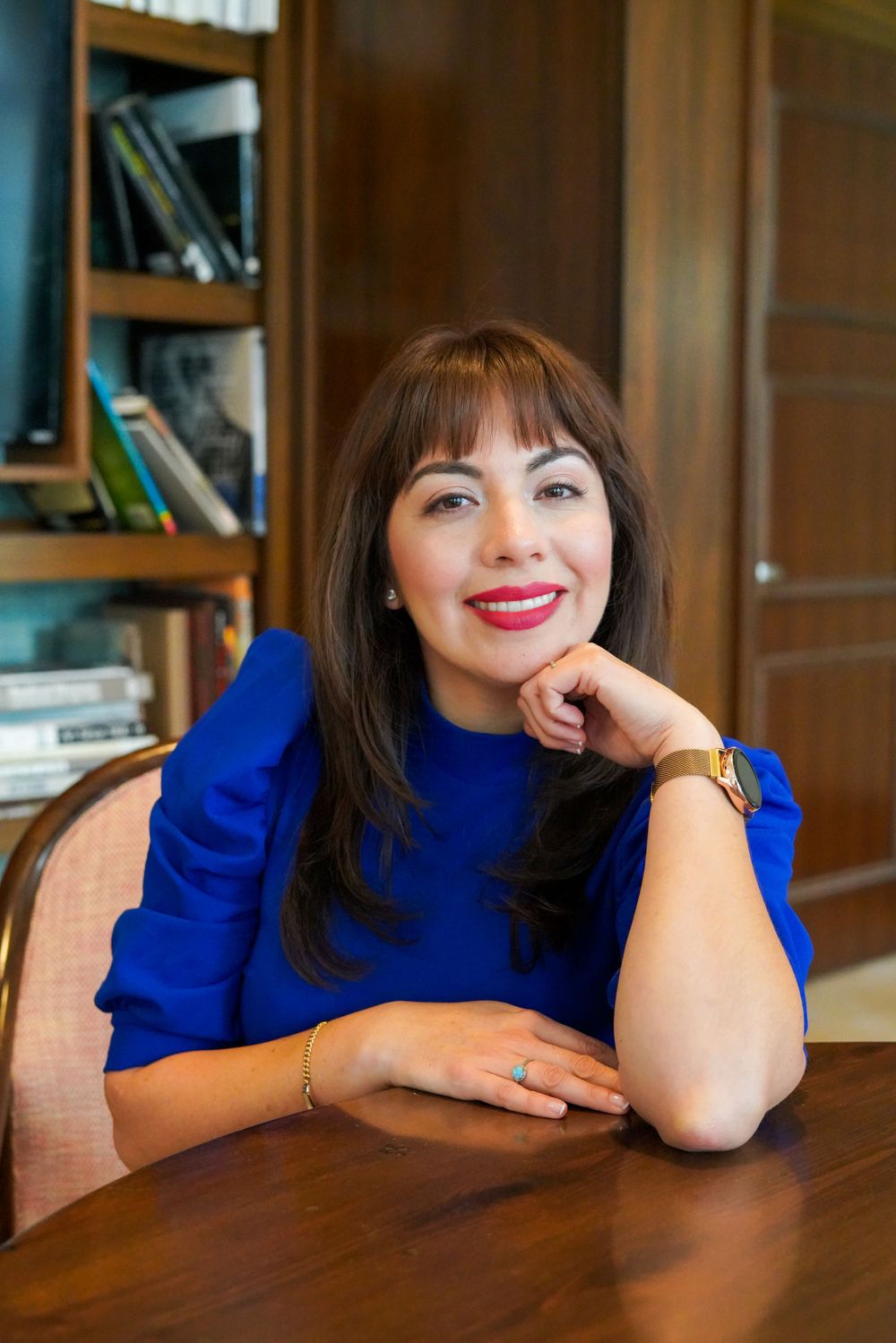 Woman in bright blue top sitting at wooden table with bookshelf background.