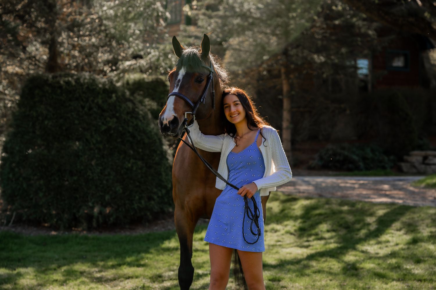 Person in blue dress poses with bay horse in front of green garden landscape.