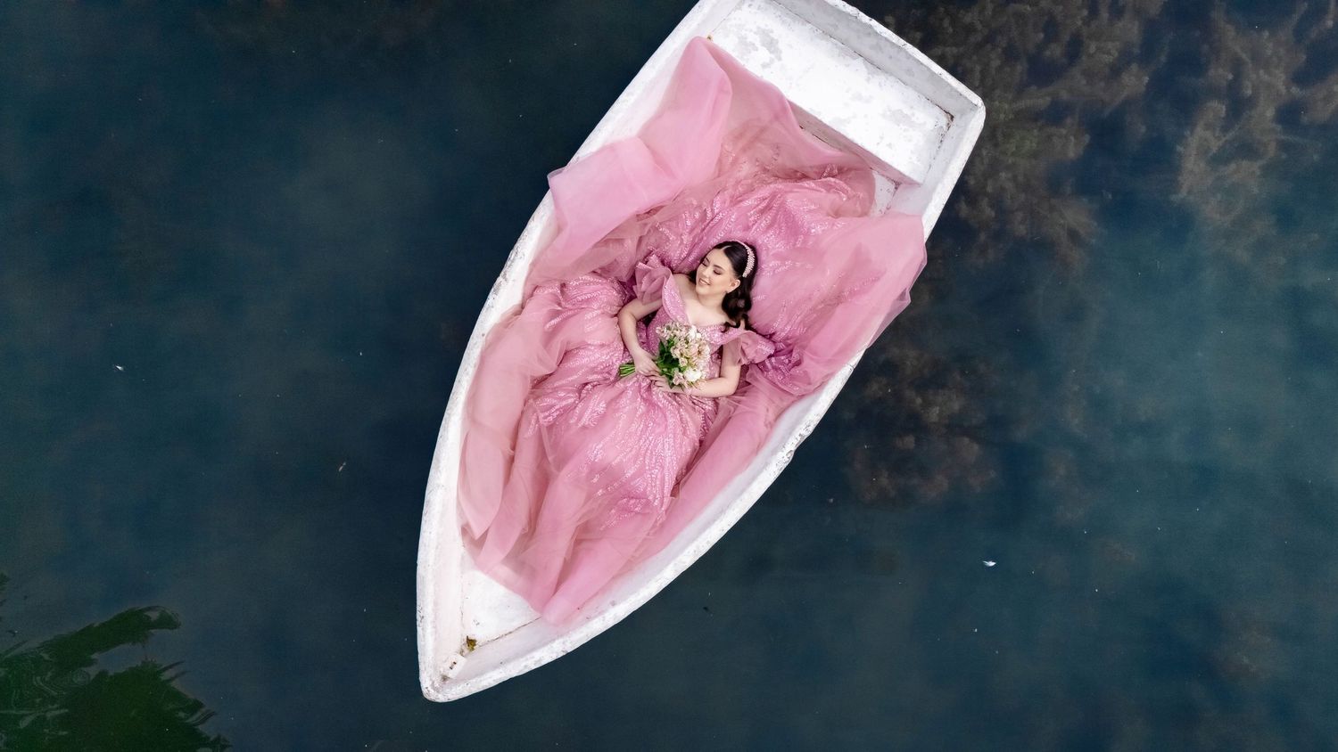 Aerial view of pink dress flowing in white boat on dark water.
