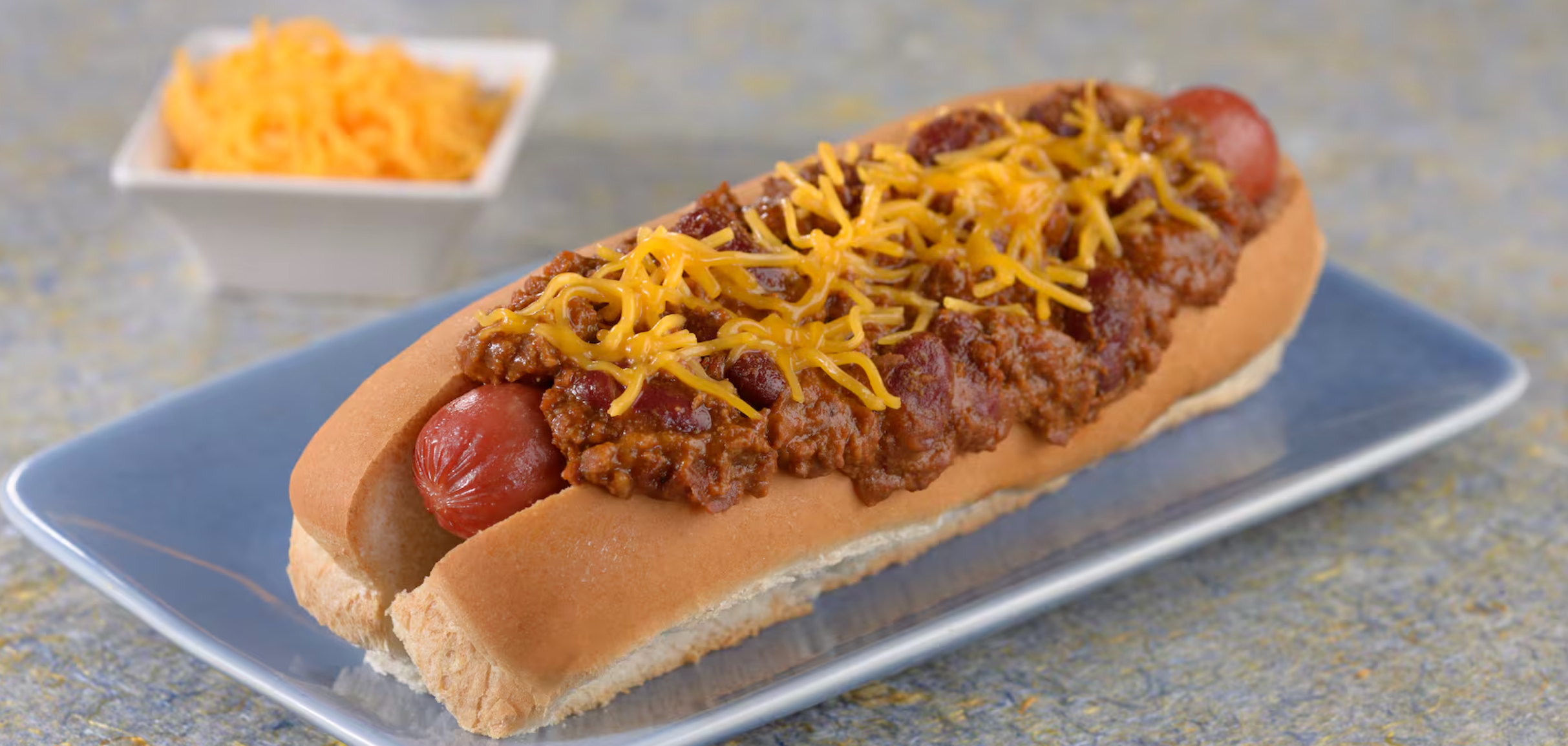 Chili cheese hot dog topped with shredded cheddar cheese and ground beef served on a blue plate.