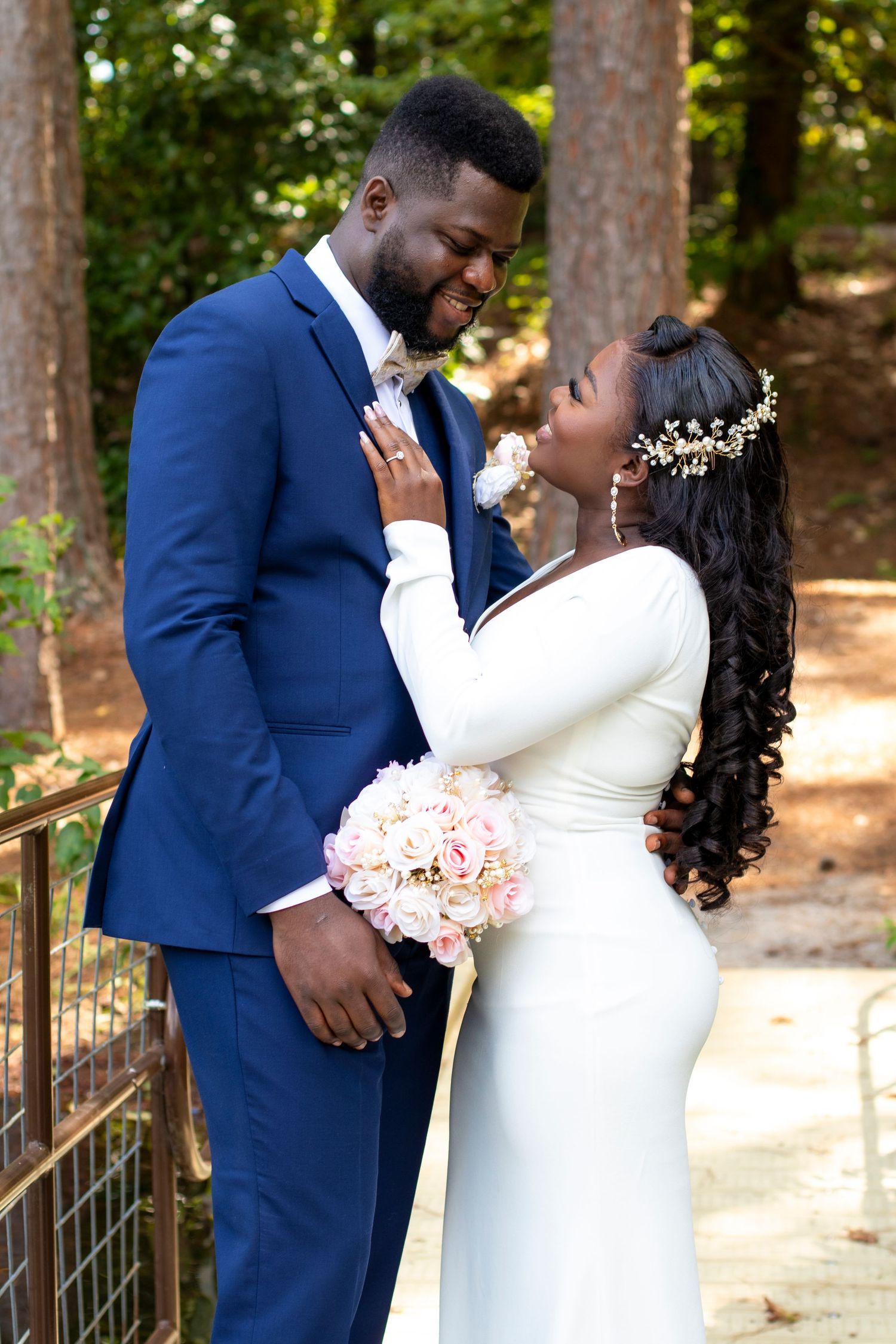 A joyful wedding couple embraces outdoors, with the bride in white holding pink flowers and groom in navy blue suit.