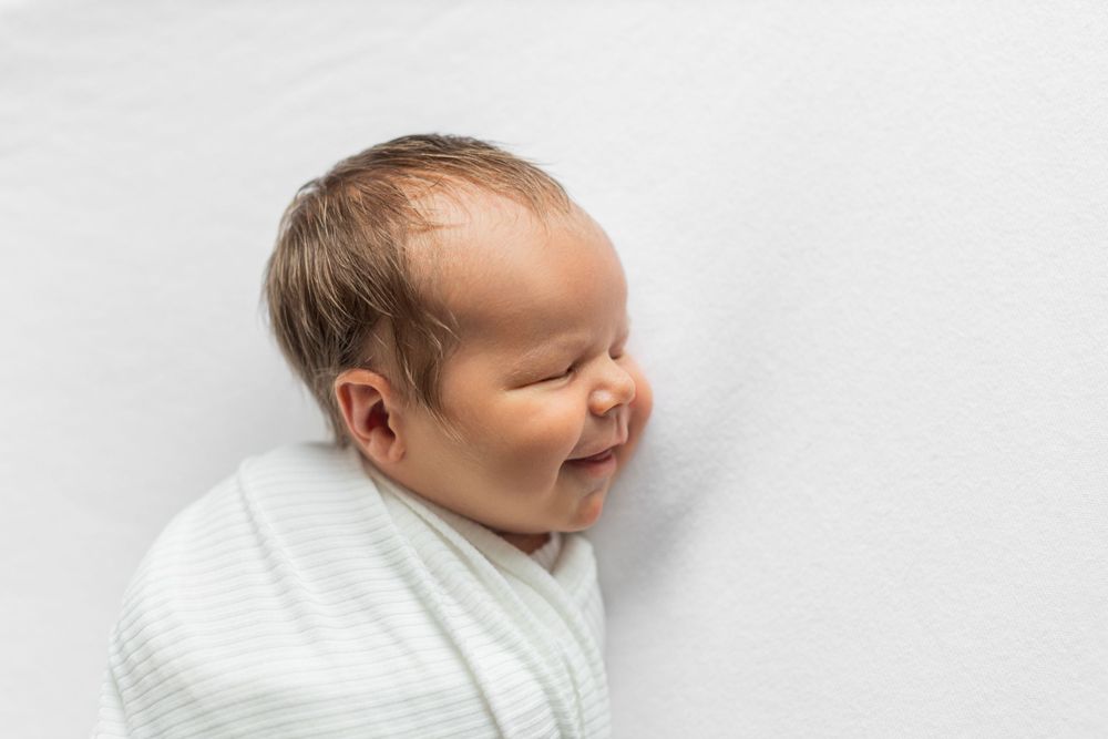 Peaceful newborn baby swaddled in white blanket smiles while sleeping against light background.