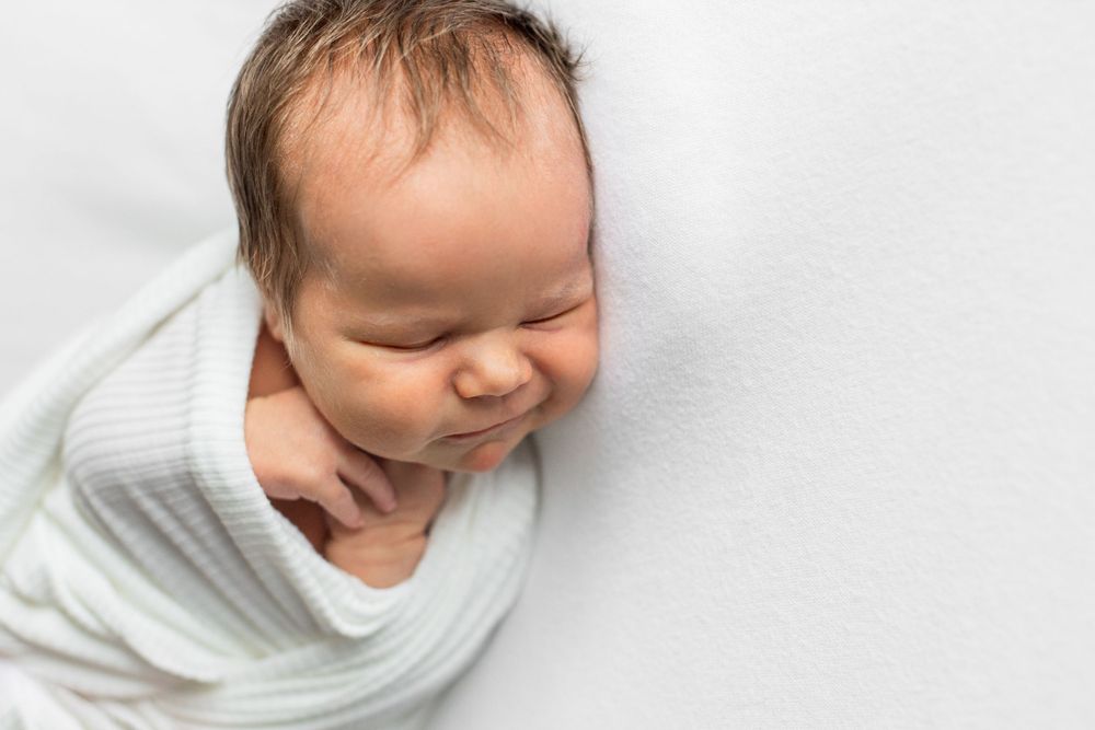 A sleeping newborn baby wearing a white knit outfit against a white background.