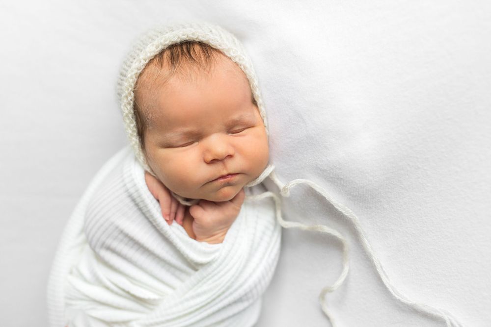 Peaceful sleeping newborn swaddled in white fabric with knit hat against white background.
