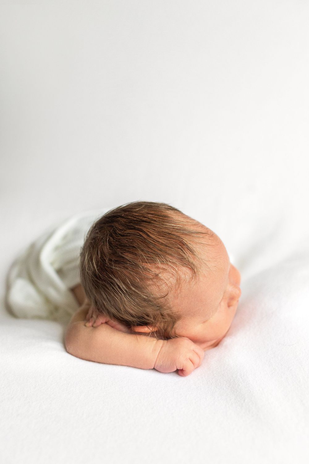 A sleeping newborn baby dressed in white rests on a white blanket.