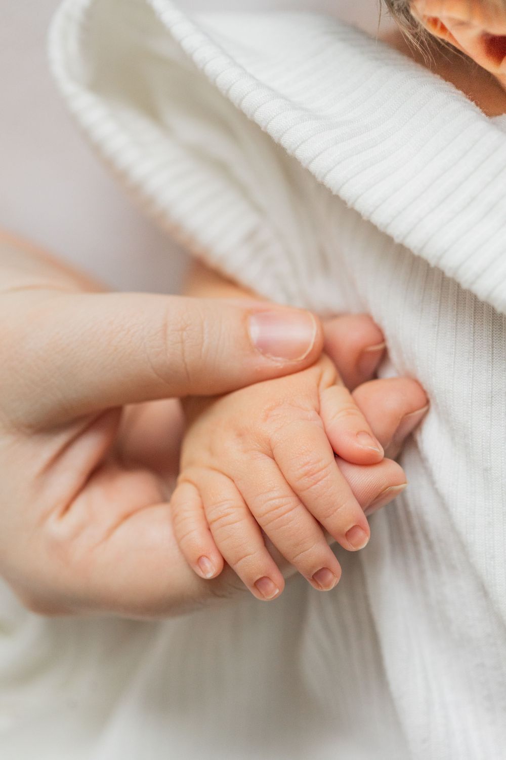 A small baby's hand gently holds an adult's finger against soft white fabric.