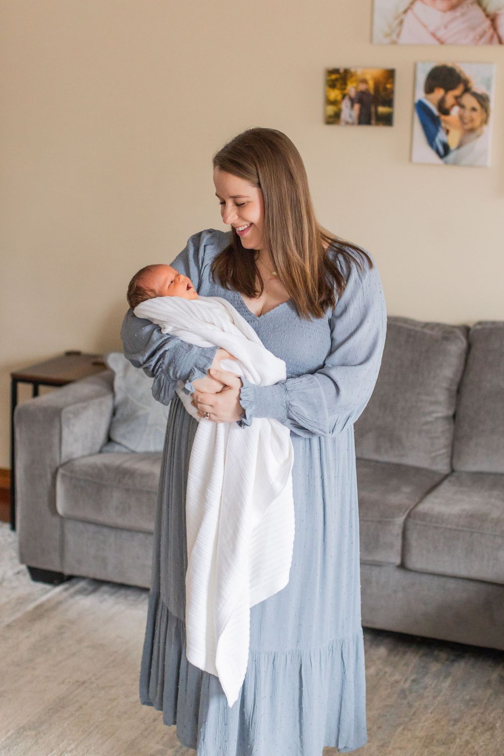 A parent in a blue dress holding a newborn baby wrapped in a white blanket in a living room setting.