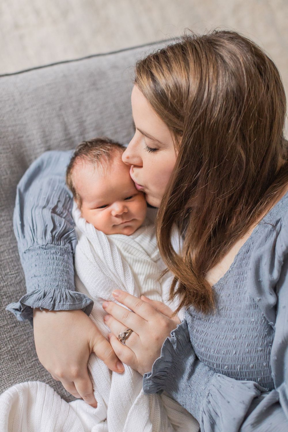 A tender moment captured as parent in blue blouse gently kisses newborn baby wrapped in white on gray couch.
