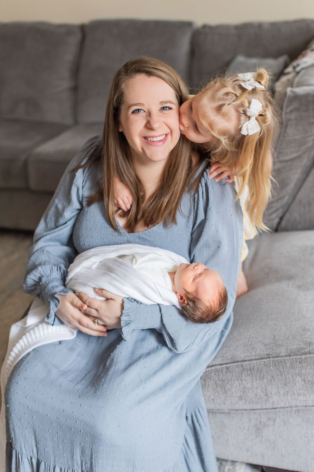 A family relaxes on a gray couch with their dog and newborn baby.