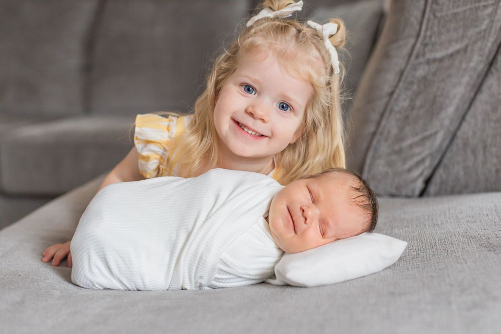 Young child lying next to newborn baby swaddled in white on gray couch cushions.