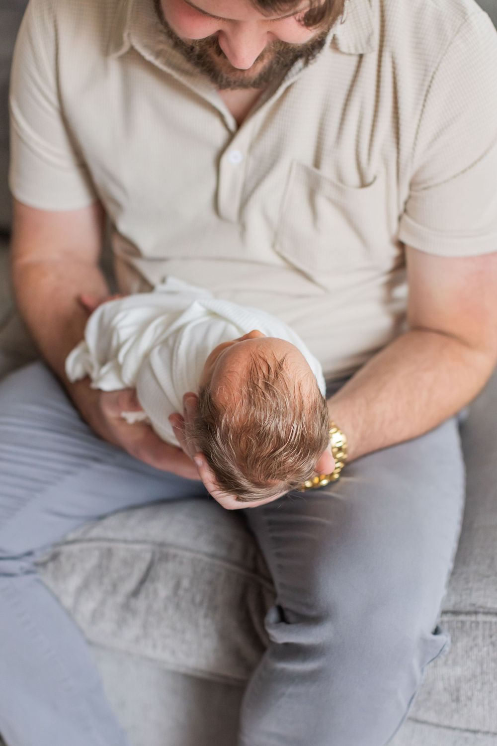 A person in a white polo shirt gazing down at a swaddled newborn baby while sitting on a couch.
