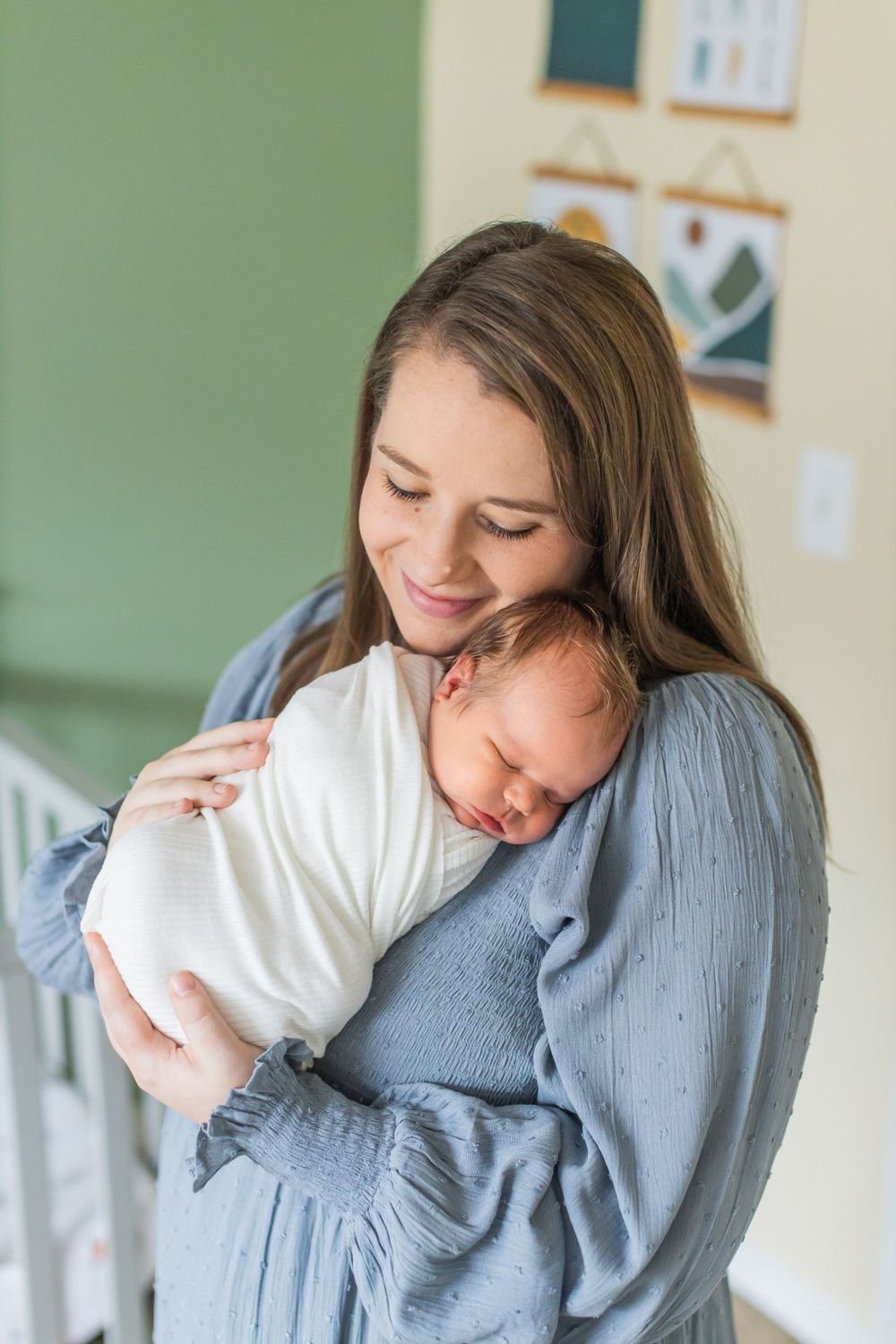 A tender moment shows a mother in a blue dress cradling her sleeping newborn baby against her chest.