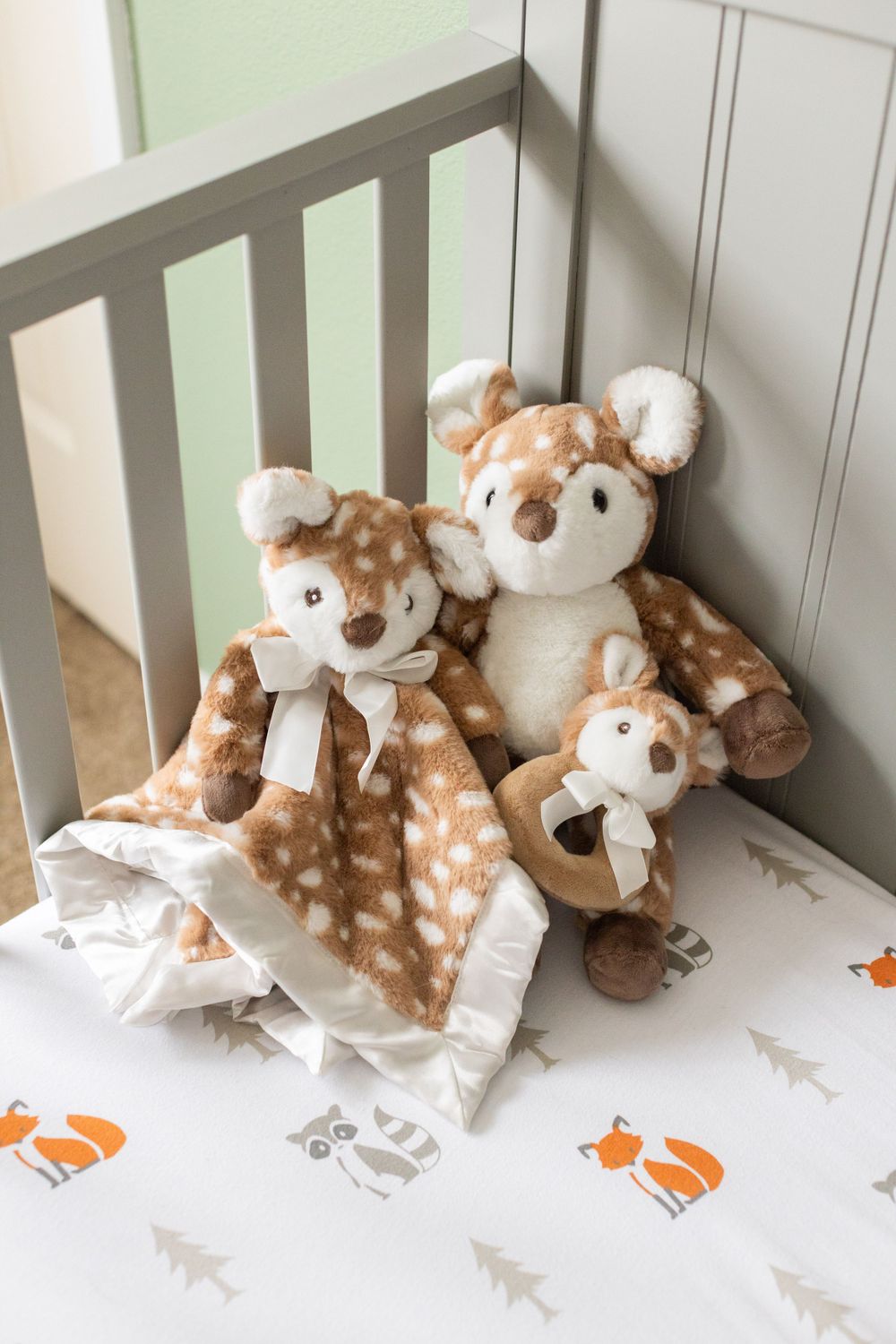 Three plush deer stuffed animals and a white blanket with fox prints sit in the corner of a gray crib.