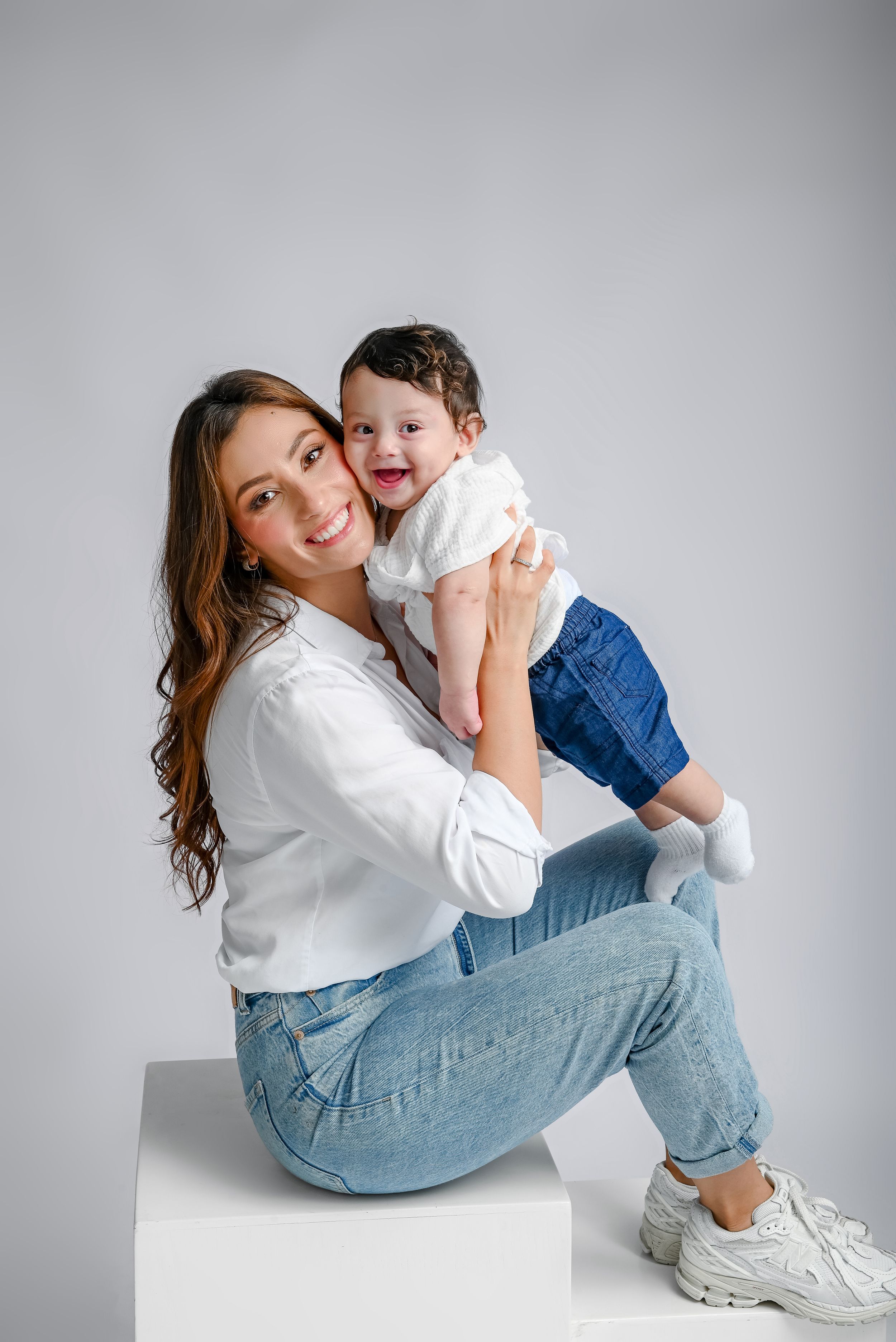 Studio portrait of parent and child in matching white tops and blue jeans sitting and hugging on white background.