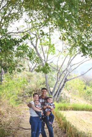 A family stands together on a dirt path surrounded by lush green trees on a sunny day.