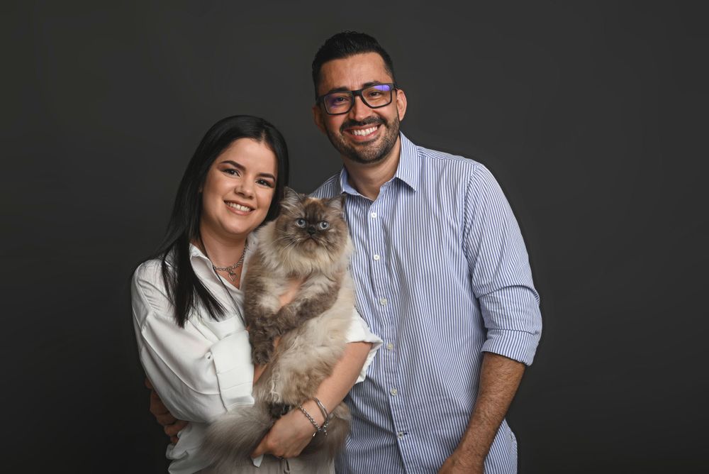 A couple in white and blue shirts holds their fluffy ragdoll cat against a dark background.