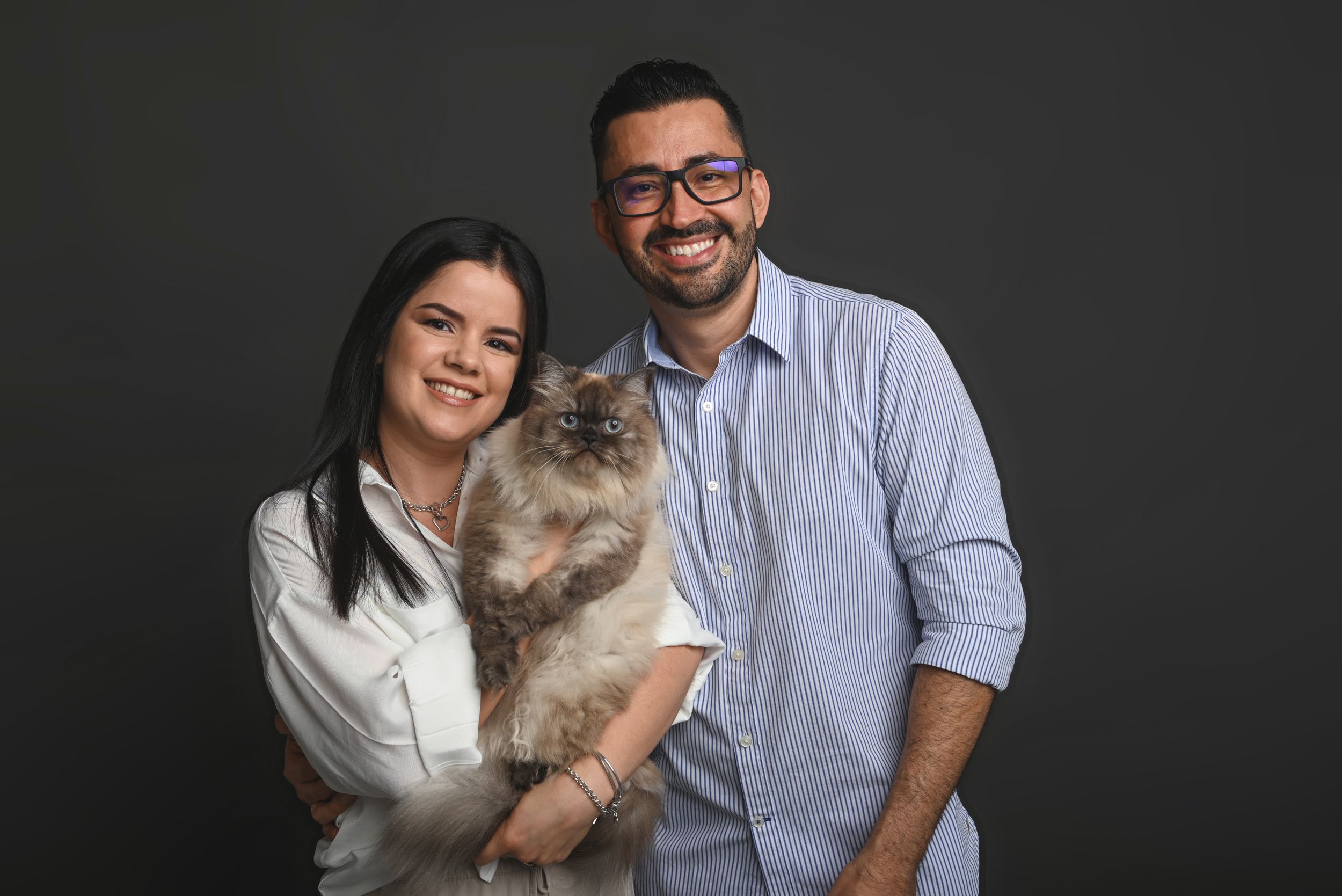 A couple in white and blue shirts holds their fluffy ragdoll cat against a dark background.