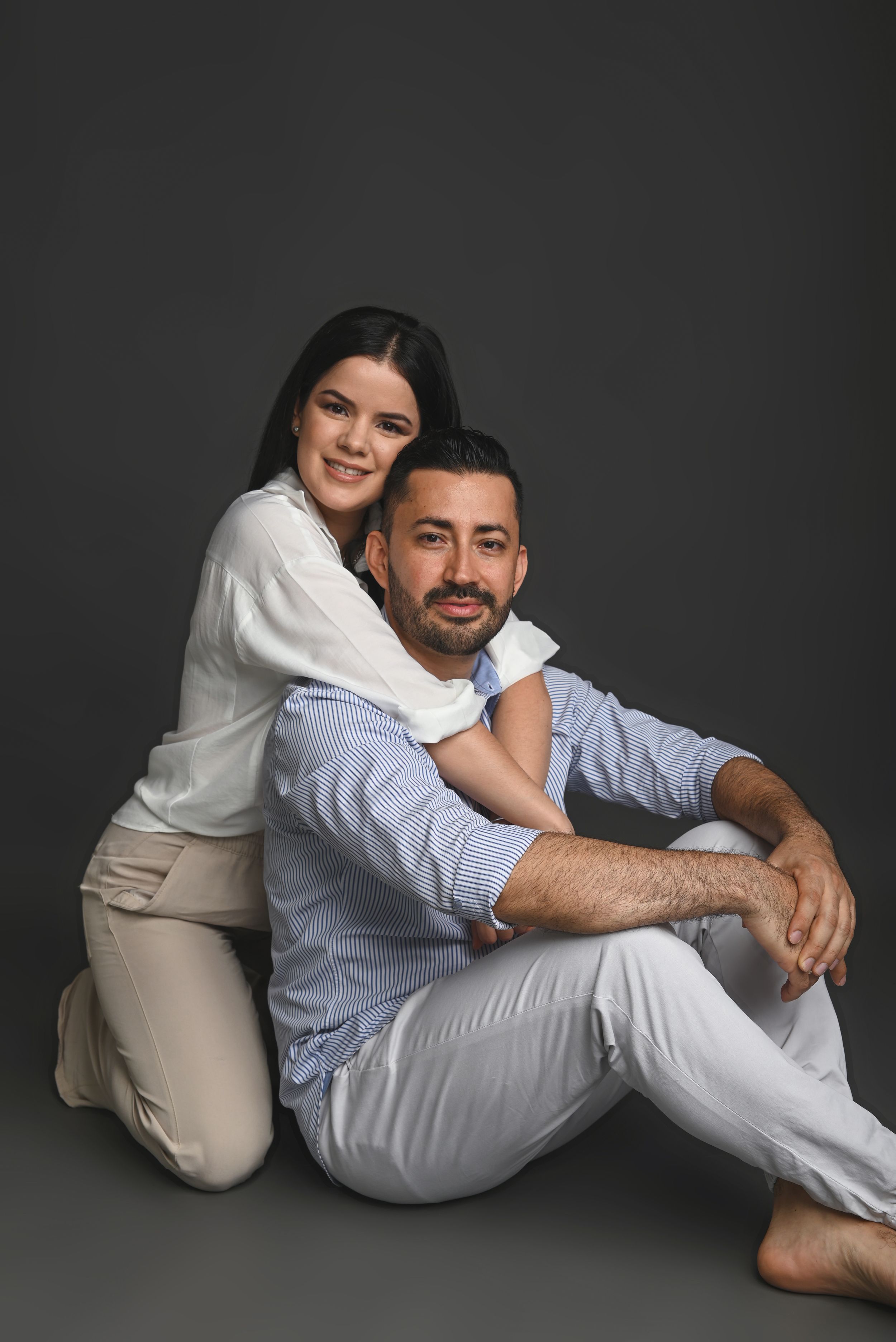 A couple in white and blue outfits pose together against dark background for an intimate portrait session.