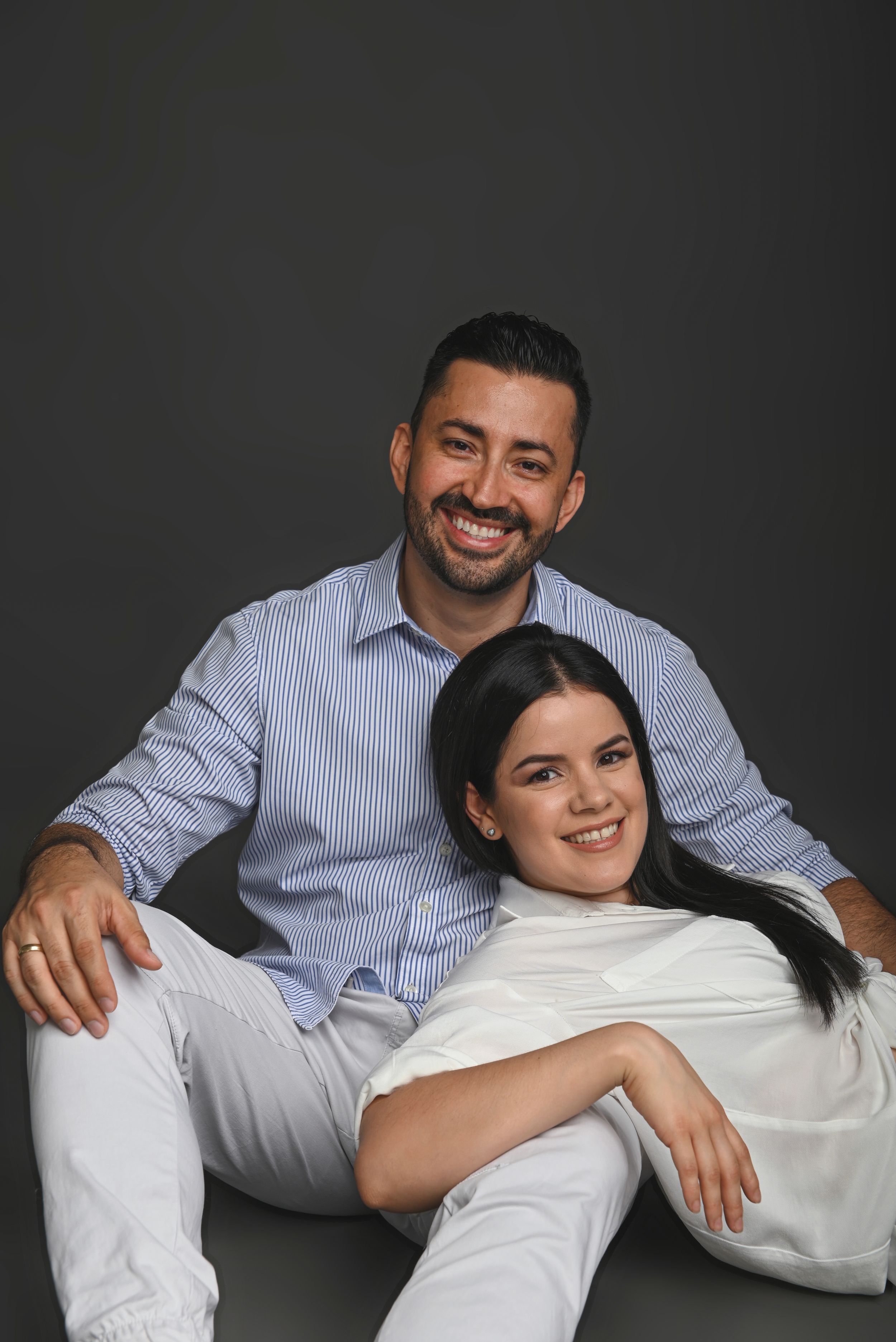 A couple in matching white and blue outfits poses together against a dark background for a portrait.
