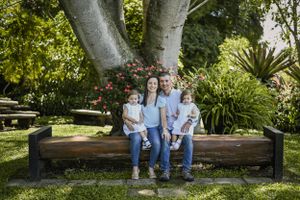 A family sits together on a wooden bench under a large tree in a lush garden setting.