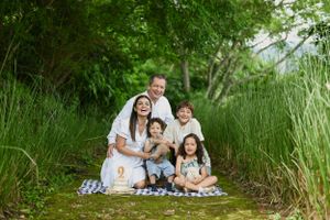 Group sitting together on picnic blanket surrounded by lush green grass and trees in outdoor nature setting.