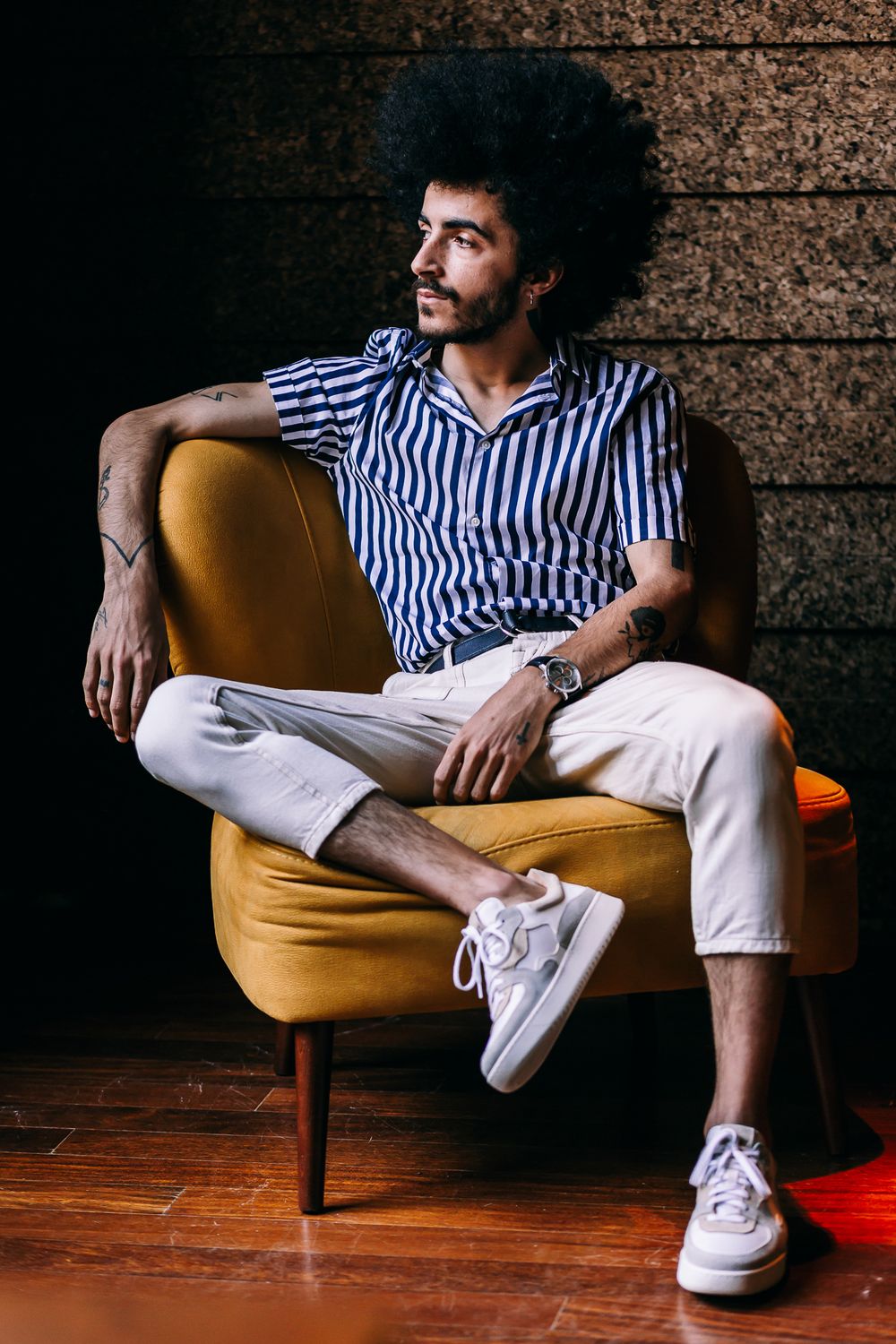 Stylishly dressed person in striped shirt and white pants lounges on mustard yellow mid-century chair against textured wall.