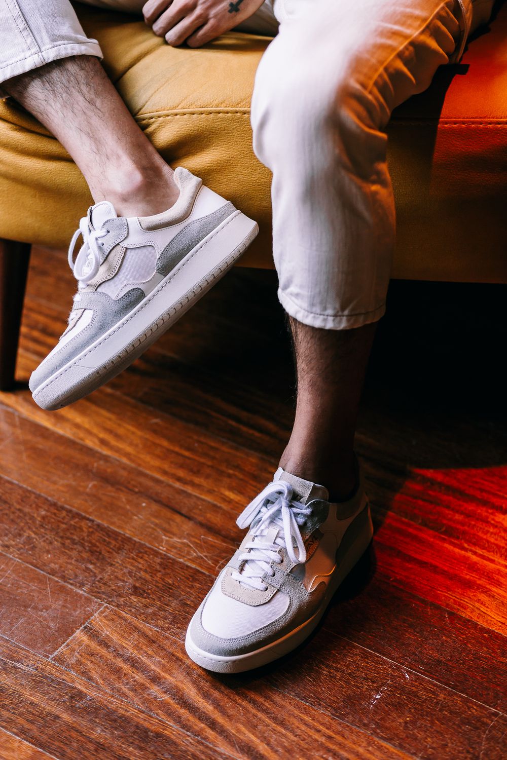 Close-up of white sneakers and cropped pants on person sitting in mustard yellow chair with wooden floor.