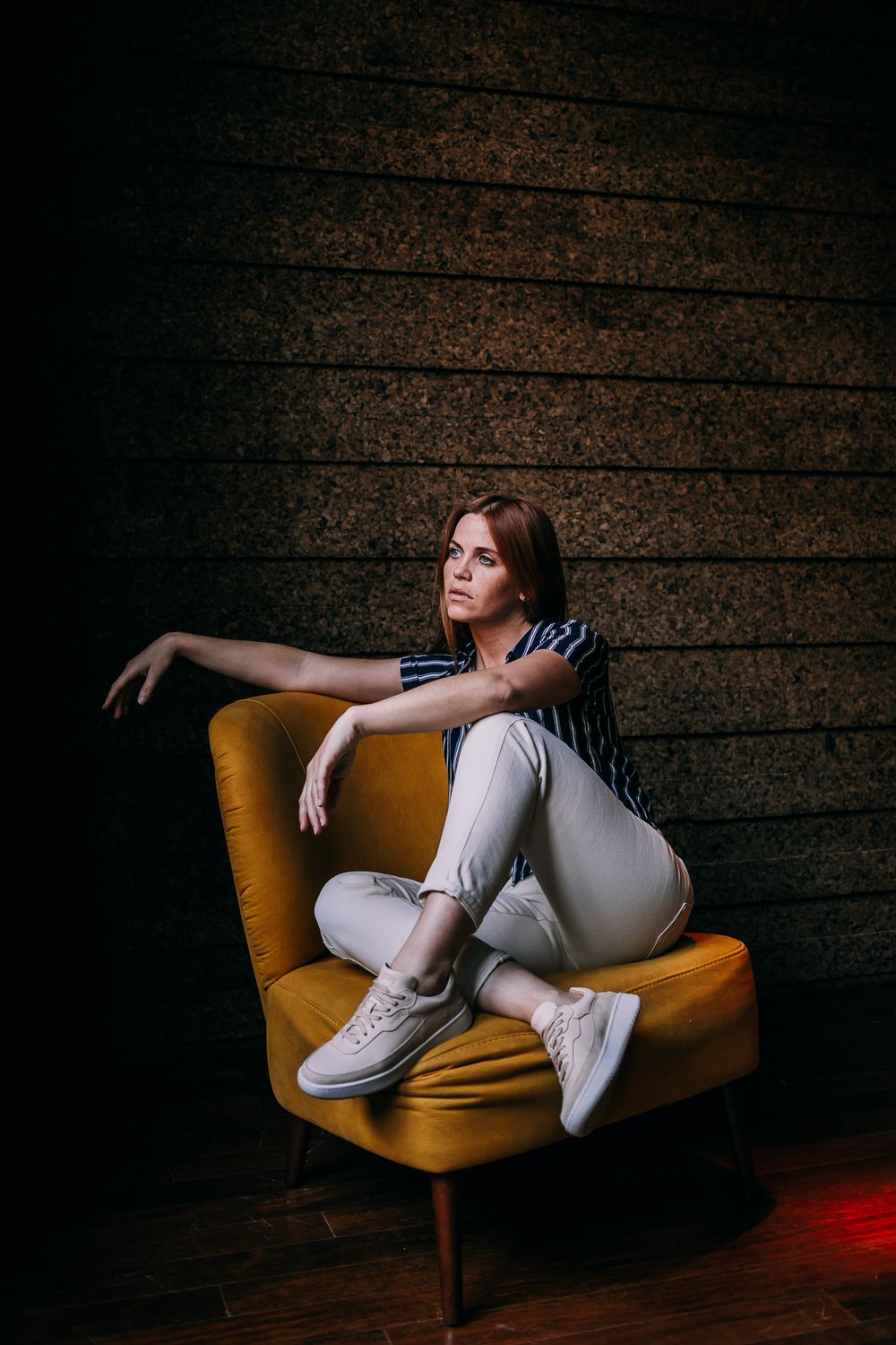 Person in casual attire lounges on vintage mustard yellow chair against dark textured wall with dramatic lighting.