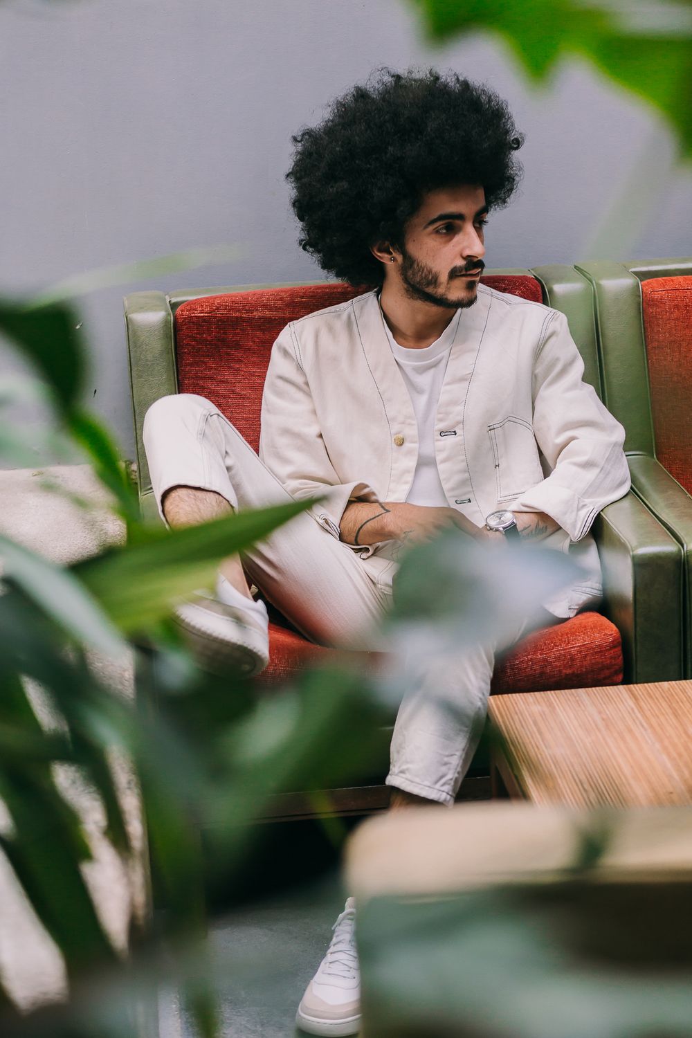 Stylish person in all-white outfit with afro hair sits contemplatively on a red couch, framed by green foliage.
