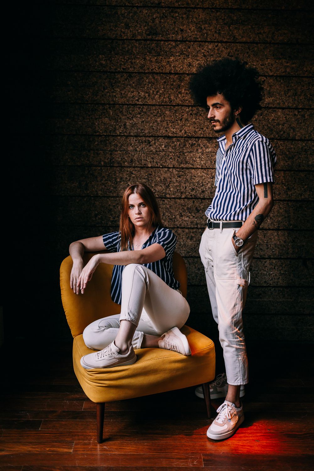 Stylish duo in striped shirts pose against brick wall with one seated in yellow chair and one standing.