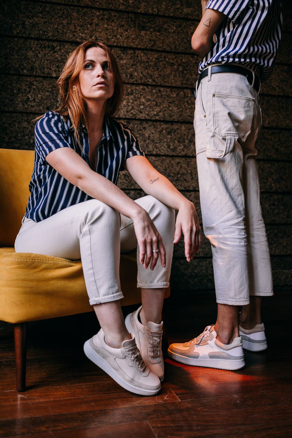 Two people in striped shirts and white pants sitting with matching sneakers on wooden floor beside yellow chair.