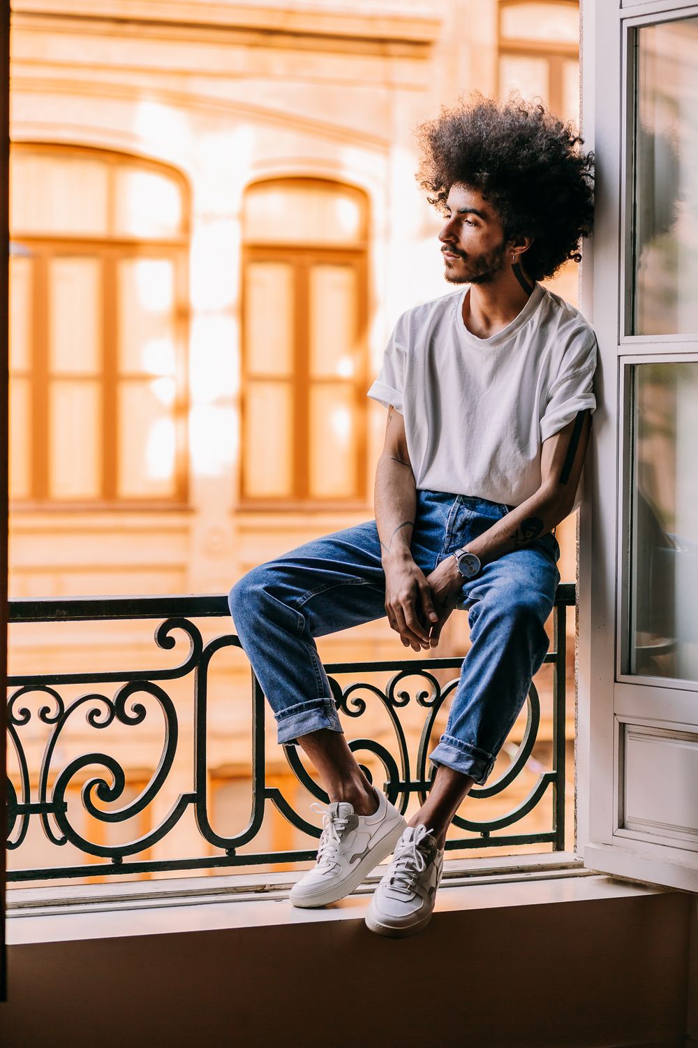 Person with curly afro sitting casually on a balcony railing wearing jeans and white t-shirt in warm sunlight.