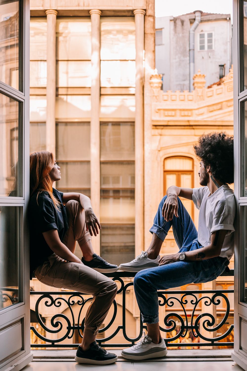 Two people sit casually on a window balcony ledge with ornate iron railings at sunset in a European-style building.