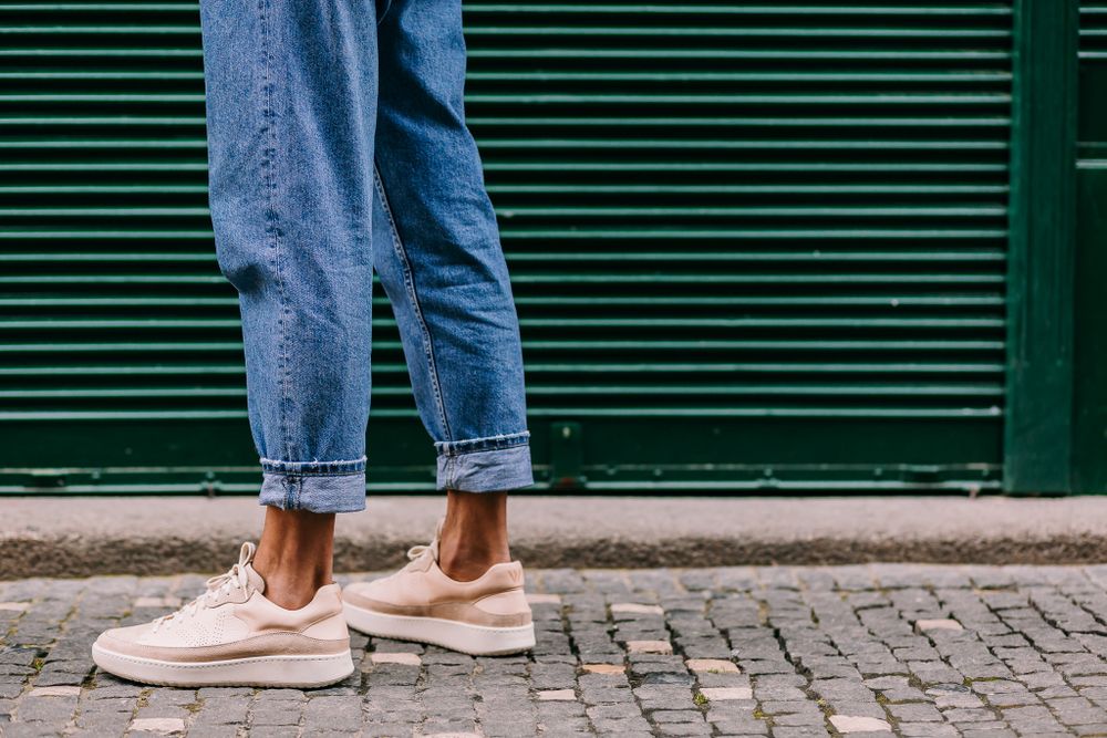 Person wearing rolled-up blue jeans and pink sneakers standing on cobblestone pavement against green shuttered wall.