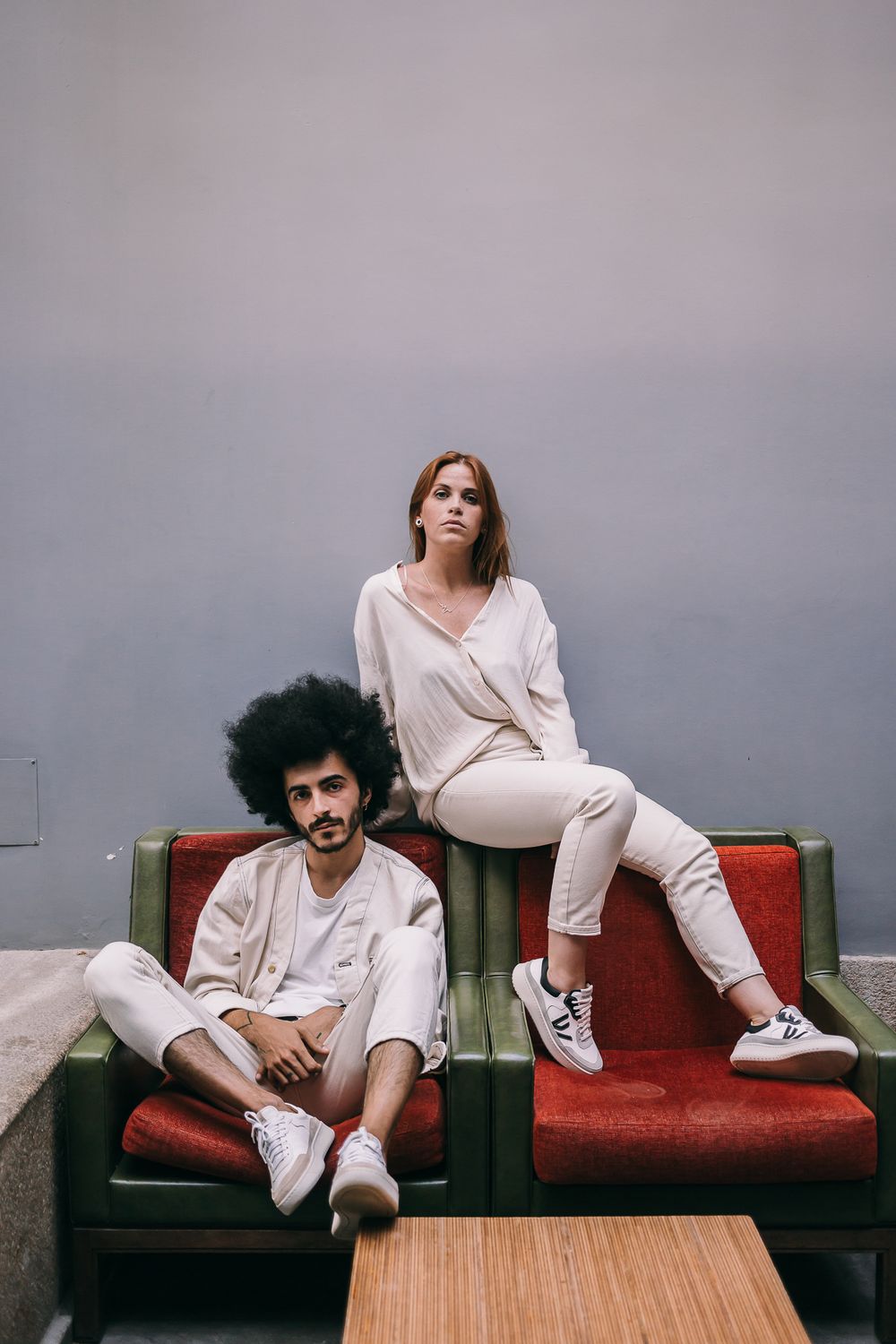Two people in casual white outfits lounging on a vintage red couch with wooden coffee table in a minimalist setting.