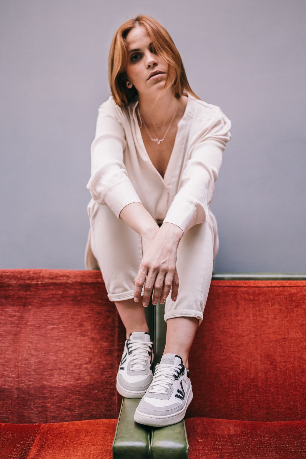 Person in monochromatic white outfit sitting casually on red upholstery wearing white sneakers against a gray backdrop.