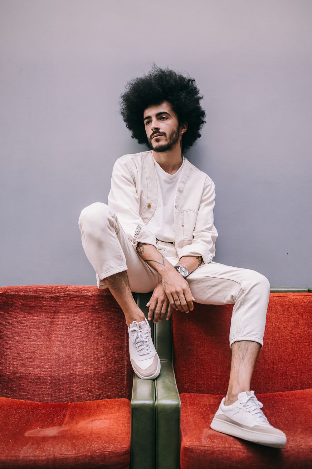 Person with natural curly hair wearing all white casual outfit sits on orange sofa against gray wall.
