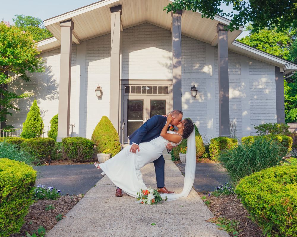 A romantic wedding couple dips for a kiss in front of a white chapel with manicured shrubs and landscaping.