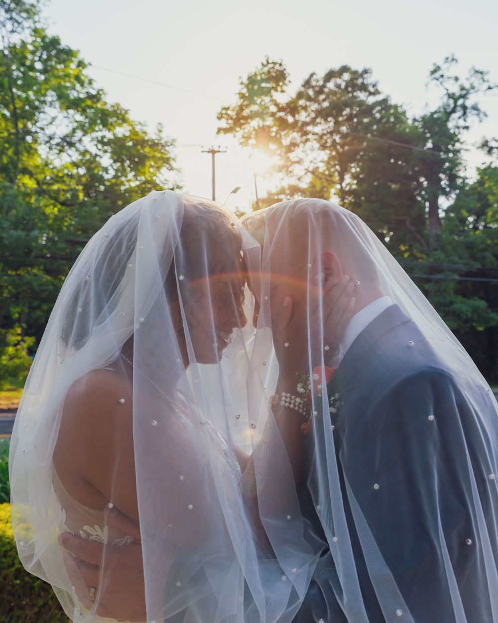 A romantic wedding couple shares an intimate moment under a flowing veil at sunset while backlit by golden sunlight.