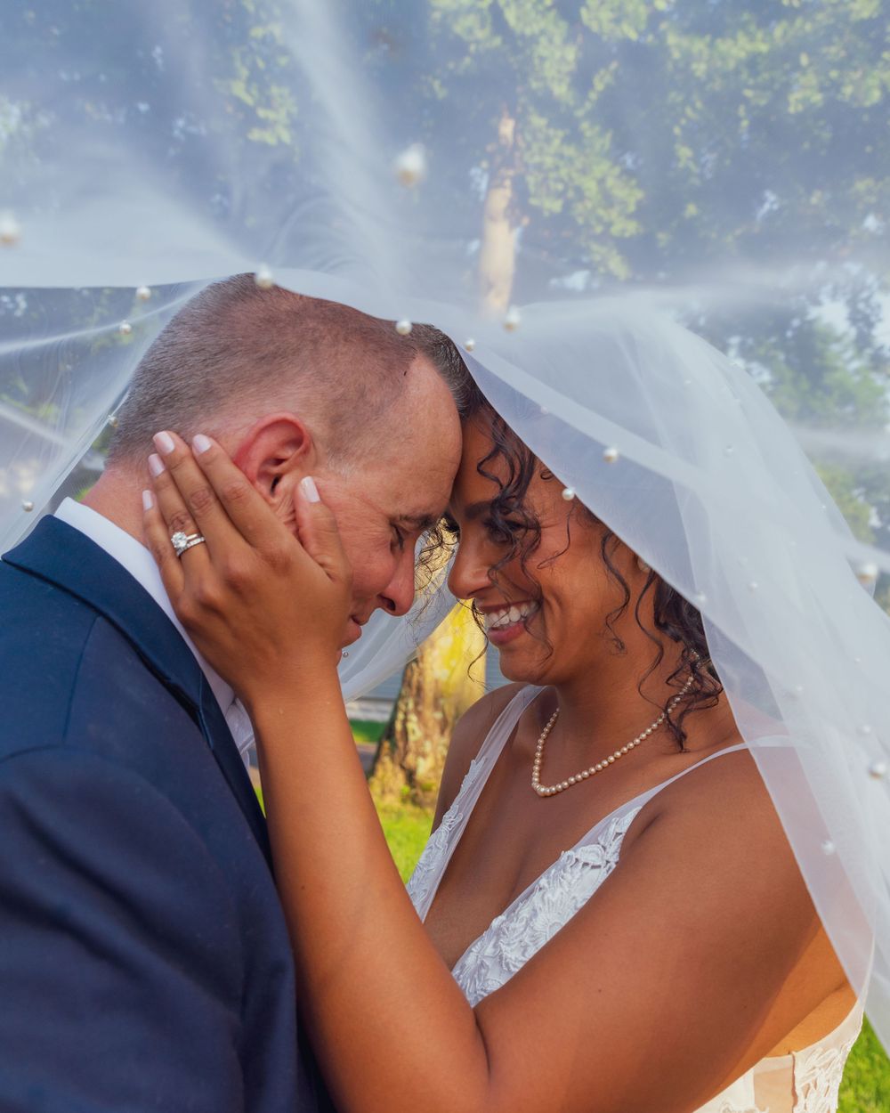 A joyful wedding couple sharing an intimate moment under a veil in front of sunlit trees on their special day.