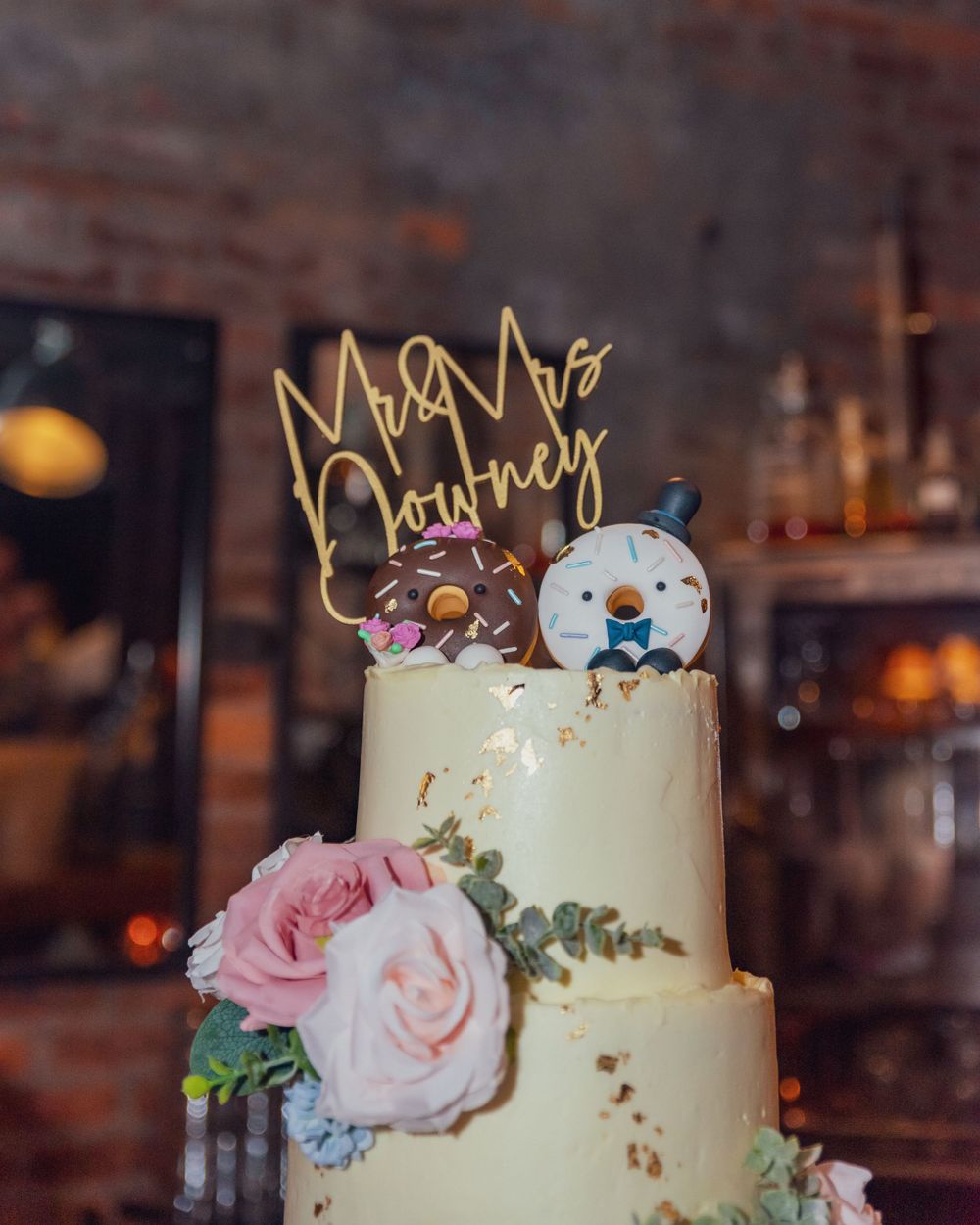 Two-tiered cream wedding cake decorated with pink roses, baby blue florals and a glowing neon 'Mr & Mrs' topper.
