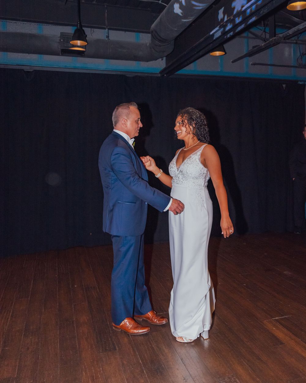 A couple shares their first dance on a dark hardwood floor, with the bride in white and groom in blue suit and brown shoes.