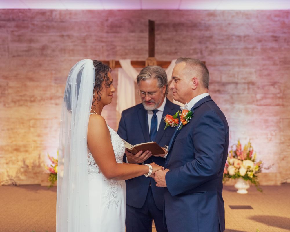 Wedding ceremony takes place in a rustic brick-walled venue with warm lighting and floral arrangements.
