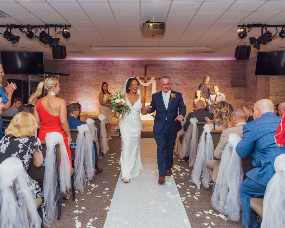 Wedding couple walks down aisle amid confetti and guests in elegant venue with brick walls and dramatic lighting.