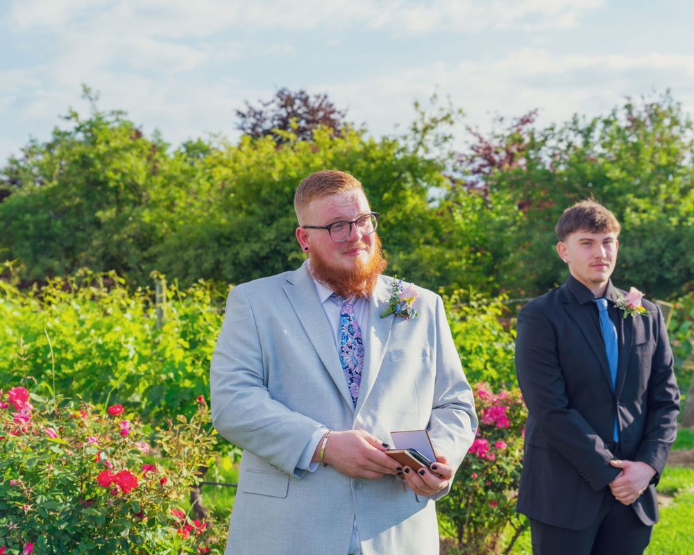 Two groomsmen in formal attire stand in a lush garden filled with colorful flowers and greenery during a wedding ceremony.