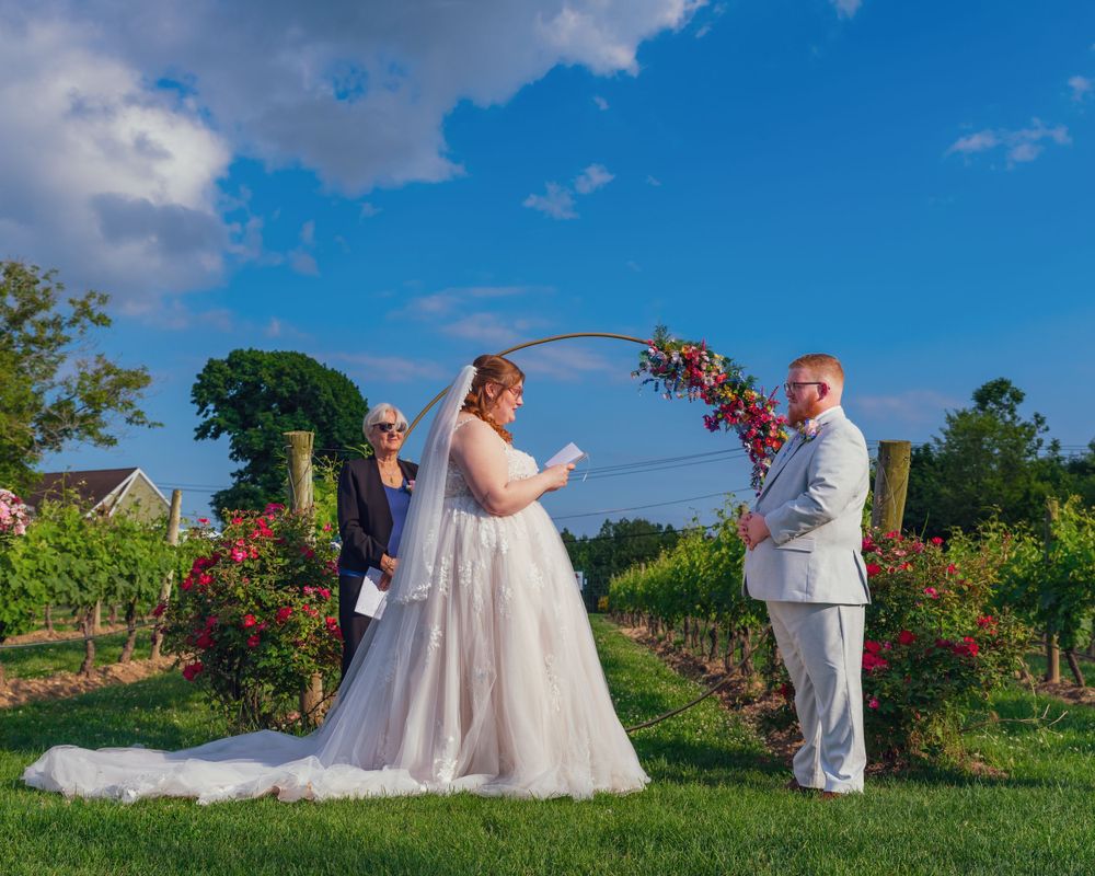 A couple exchanges vows under a floral arch during their outdoor vineyard wedding ceremony on a beautiful summer day.