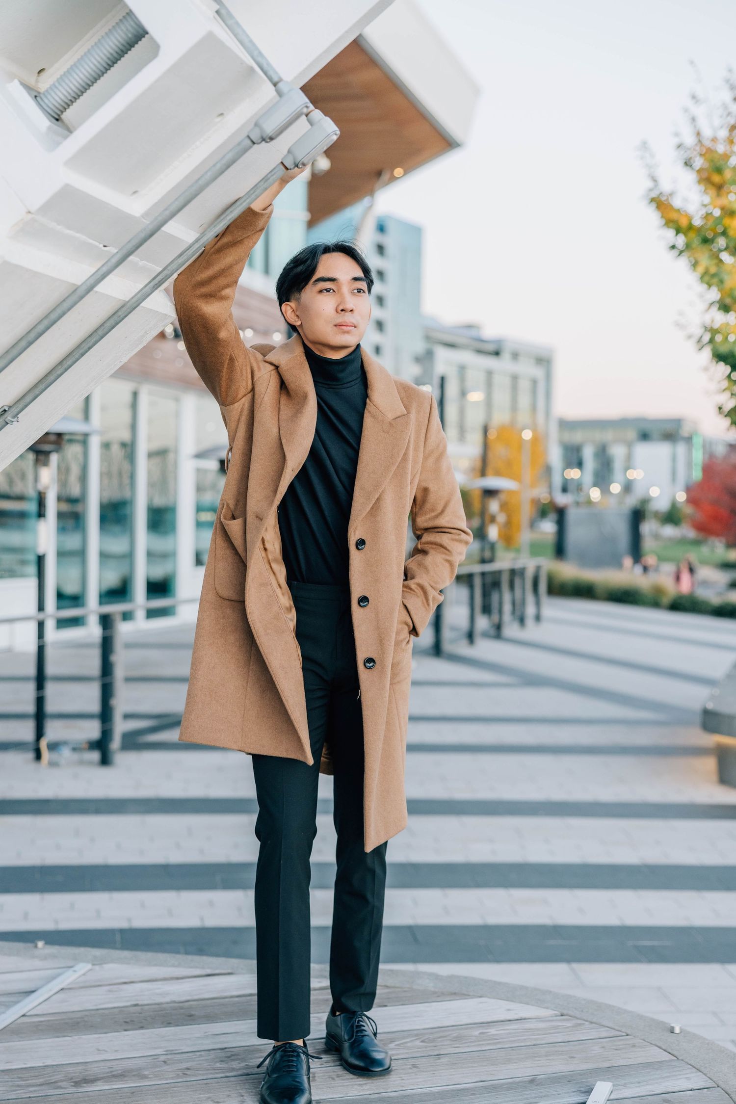 Stylish young man in a camel overcoat and black turtleneck posing outdoors in an urban setting.