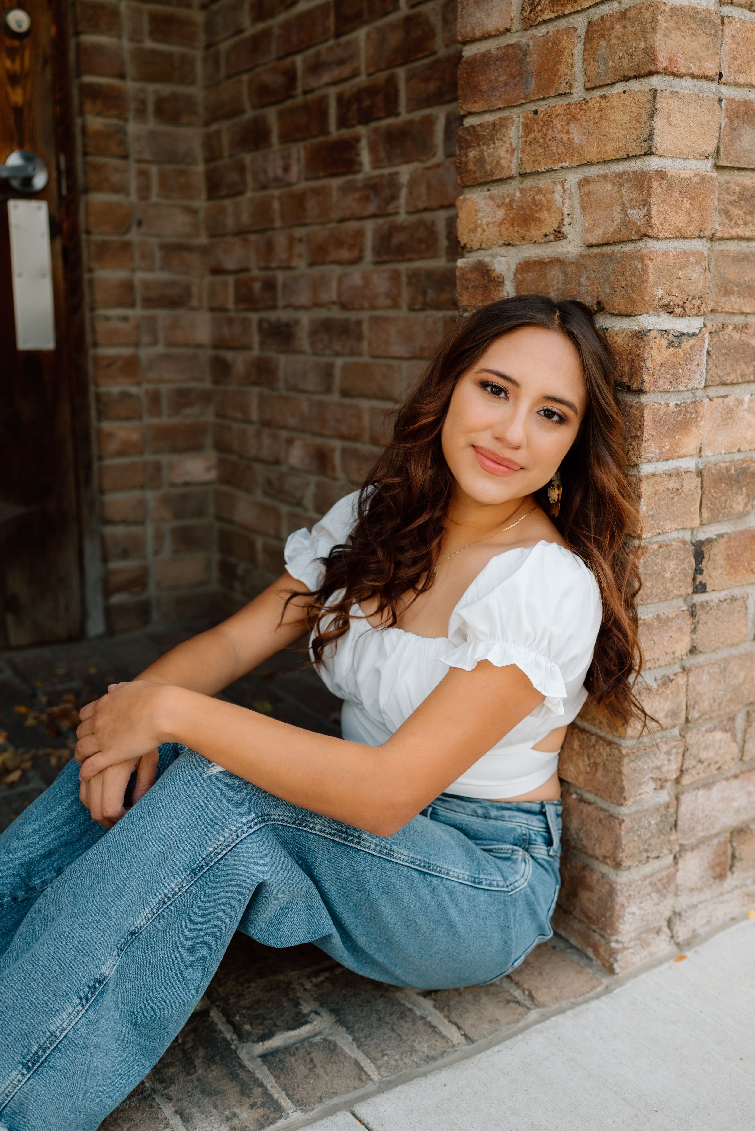 Young woman in white peasant top and blue jeans sits casually against brick wall in natural outdoor lighting.