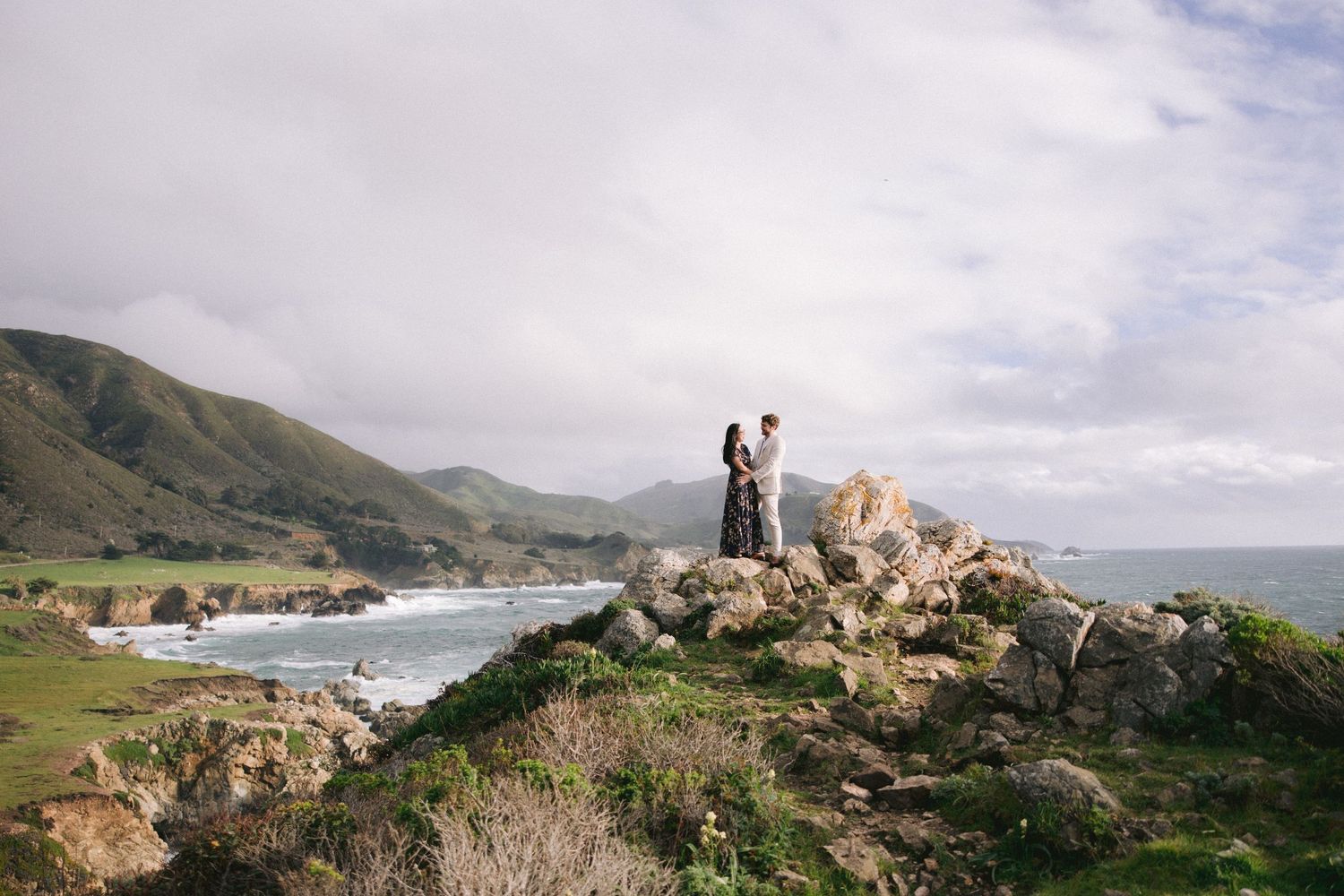 Two people stand atop coastal cliffs overlooking a scenic bay and mountains at Big Sur, California.