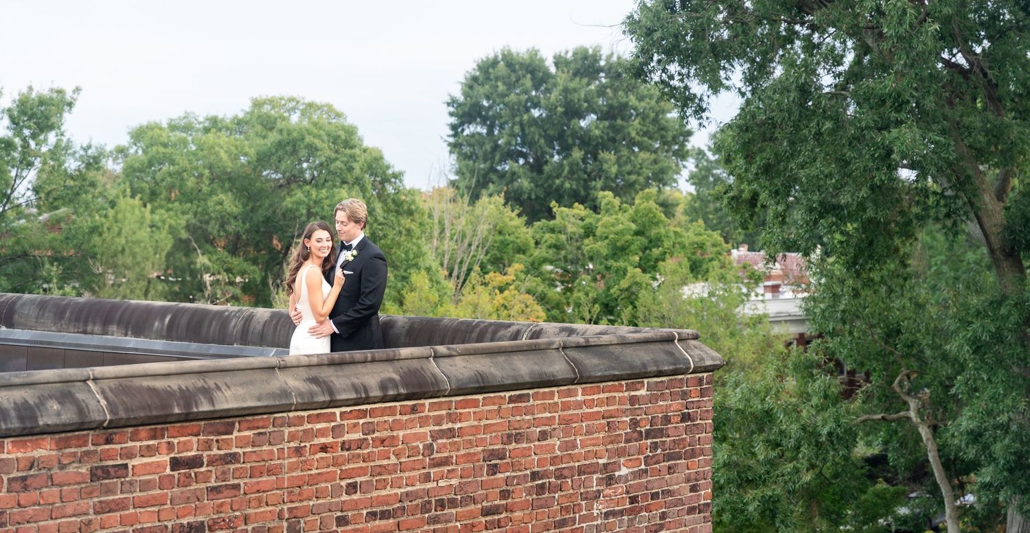 Bride and groom embracing on a brick rooftop wall surrounded by lush green trees.